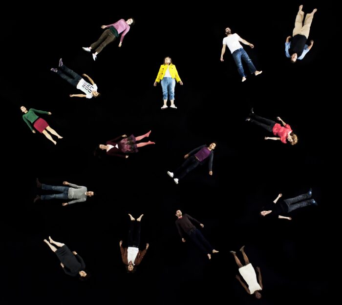 Fotografia. Ujęcie z góry. Na czarnej podłodze leży kilkanaście osób w kolorowych ubraniach. Leżą na wznak z rękoma wyciągniętymi wzdłuż ciała.
