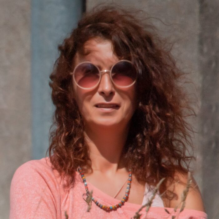 A woman with a storm of curly hair, wearing round sunglasses. She is standing somewhere outdoors, wearing a pink blouse, coloured beads around her neck.