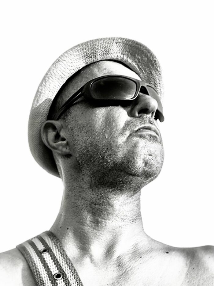 Black and white photo of a man looking up. He is weraing a summer hat and sunglasses, a probable stripe of a drum over his shoulder.