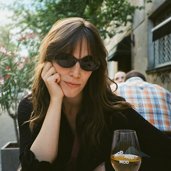 A young woman sitting at the table somewher in the streets of the city. She has long dark hair and sunglasses. She supports her head with her hand. The supportng elbow resting over the table.