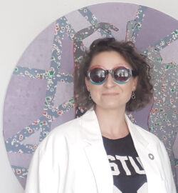 Portrait photograph. A woman with dark curly hair, wearing sunglasses and a white jacket stands against a purple circle graphic.