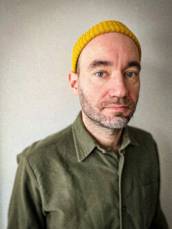 Zdjęcie mężczyzny w średnim wieku z lekkim zarostem, w koszuli w kolorze khaki i żółtej czapce.