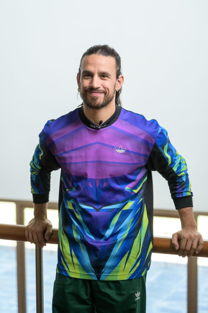 Portrait of Marco da Silva Ferreira. He is standing in a colorful sweatshirt, looking at the camera, with dark hair tied in a ponytail and a beard and mustache. He is smiling.