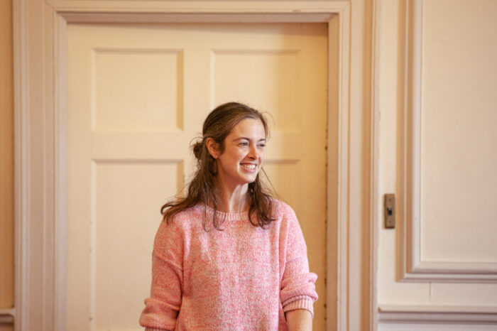 Zdjęcie portretowe Amy Draper. Długowłosa młoda uśmiechnięta szatynka w różowym swetrze.