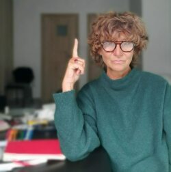 Portrait of Bogna Świątkowska - a woman with fair culrly hair and glasses wearing a green jumper and pointing her finger up